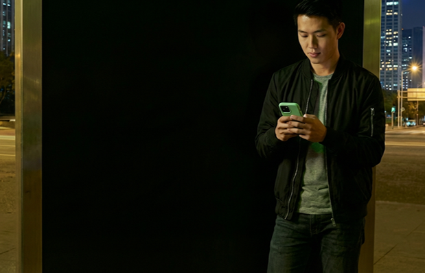 Uma mulher negra de cabelos raspados e descoloridos segura um smartphone com as duas mãos. Ela está em um ambiente urbano noturno e atrás dela passa um carro verde neon.